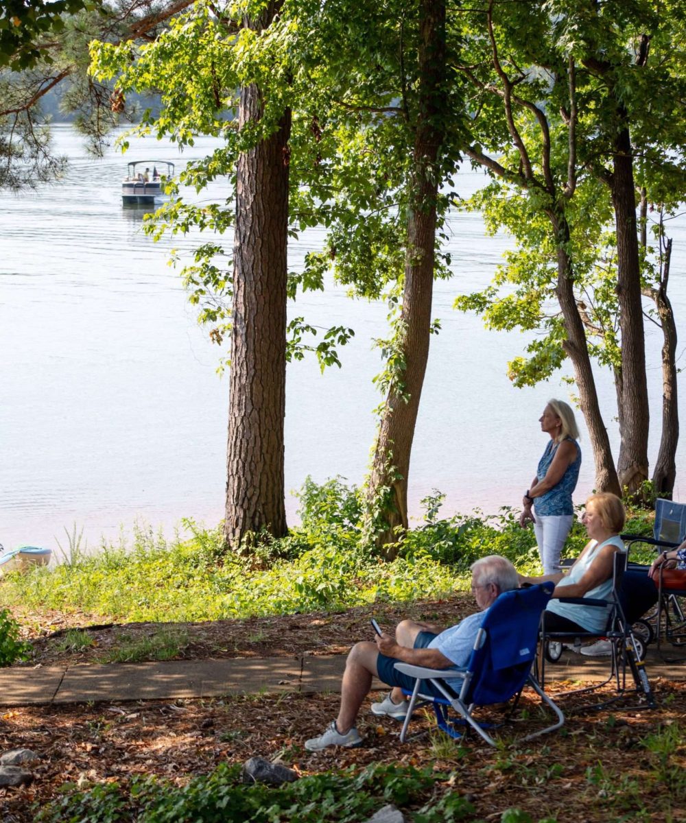 people sitting and standing on the edge of west point lake worshipping at church on the hooch with a boat in the distance